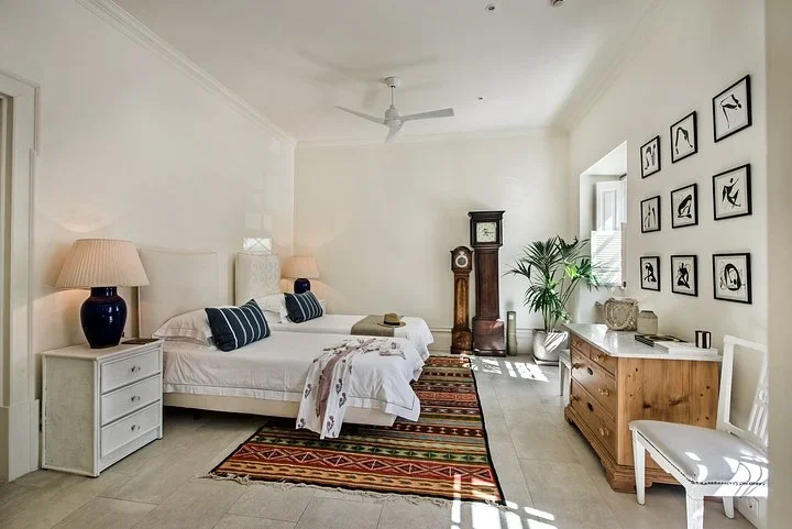 Bright bedroom with two twin beds, a striped rug, a grandfather clock, and framed line drawings on the wall