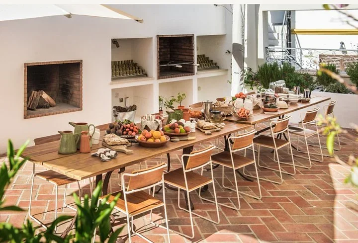Long wooden table in a brick courtyard set with fruit bowls, bread, jugs, and coffee cups for breakfast