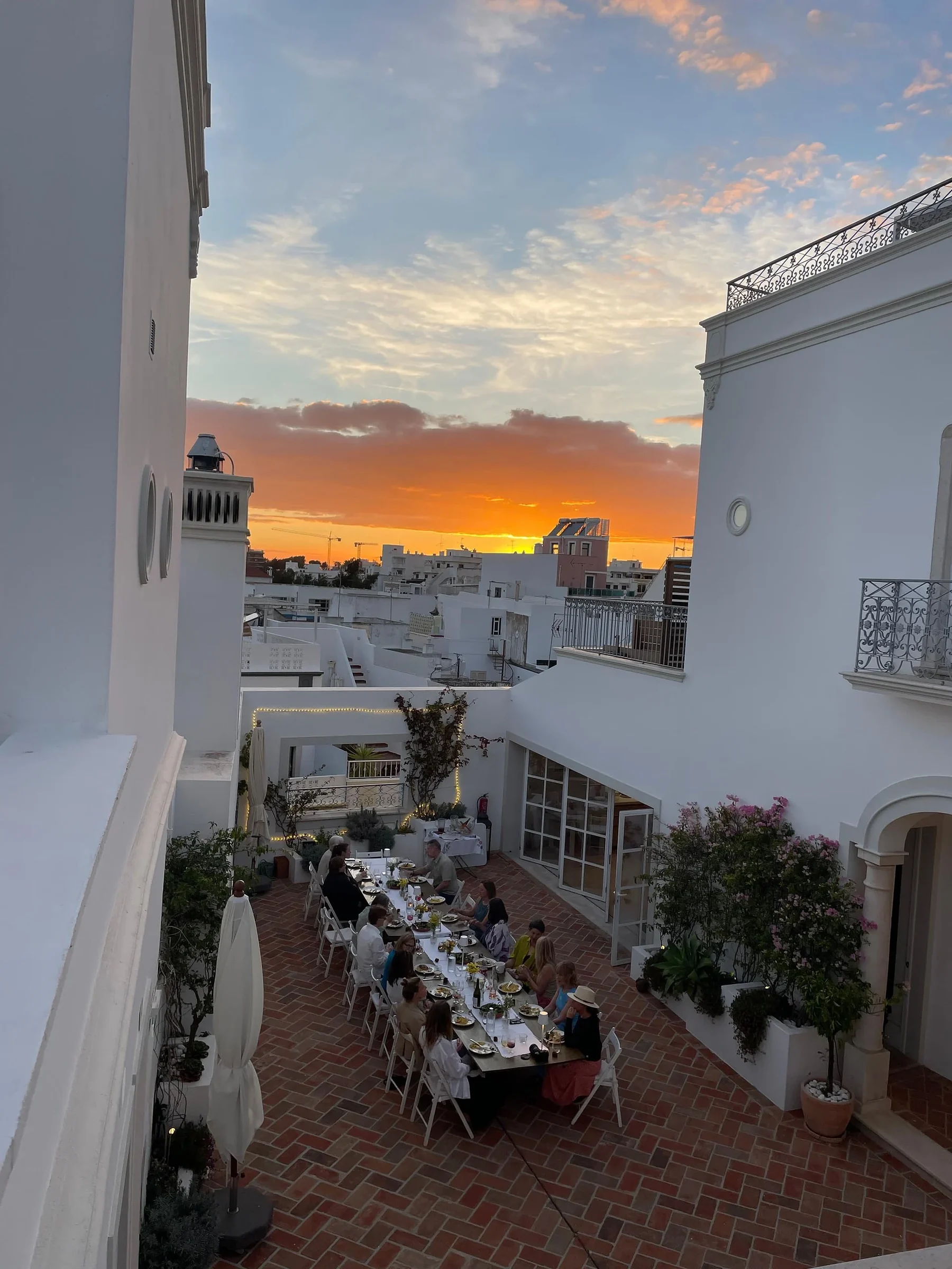 Guests sharing dinner at a long table in a white courtyard with bougainvillea, as an orange sunset lights the rooftops