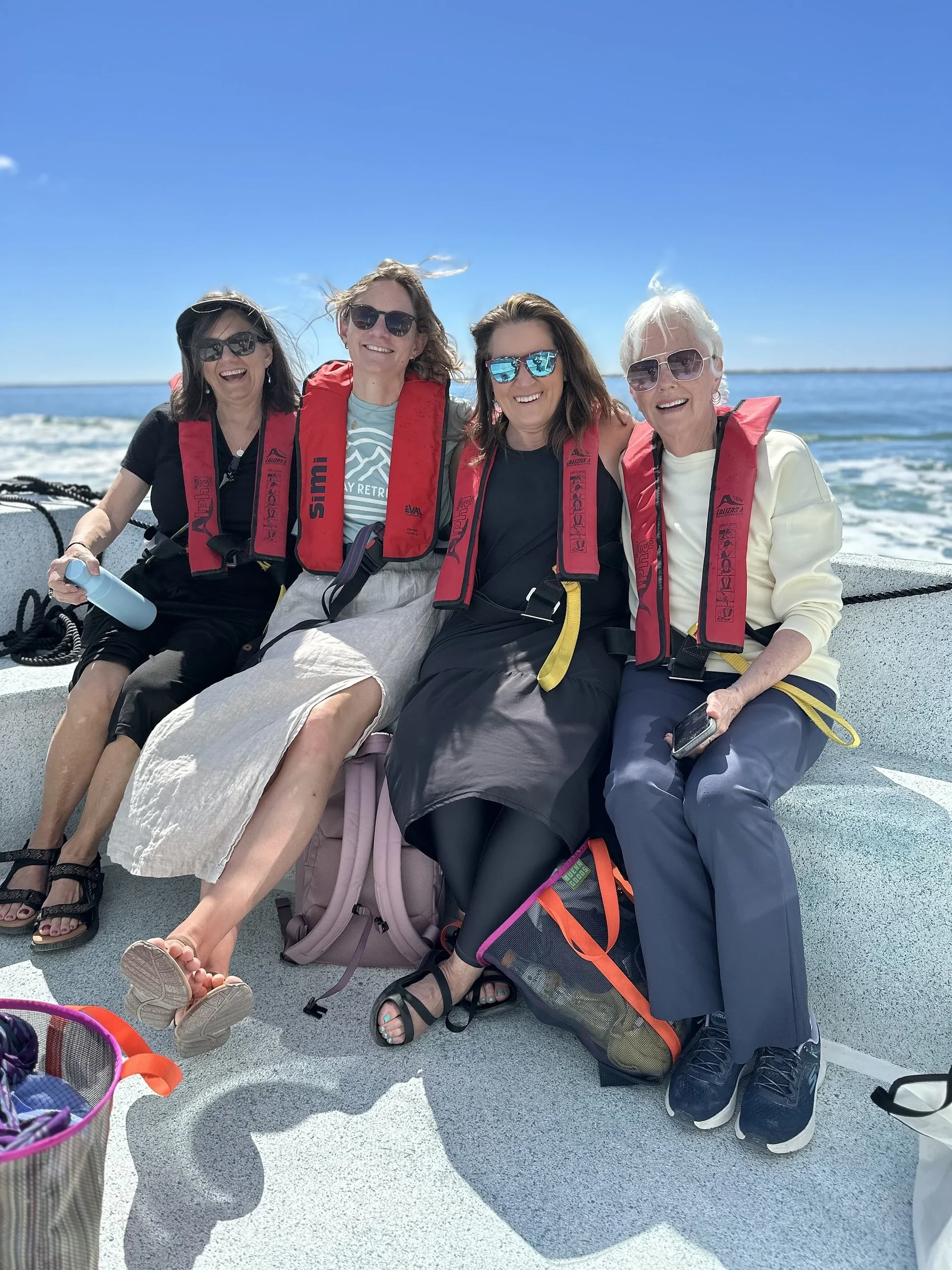 Four smiling women in red life jackets sitting together at the back of a boat on a sunny day