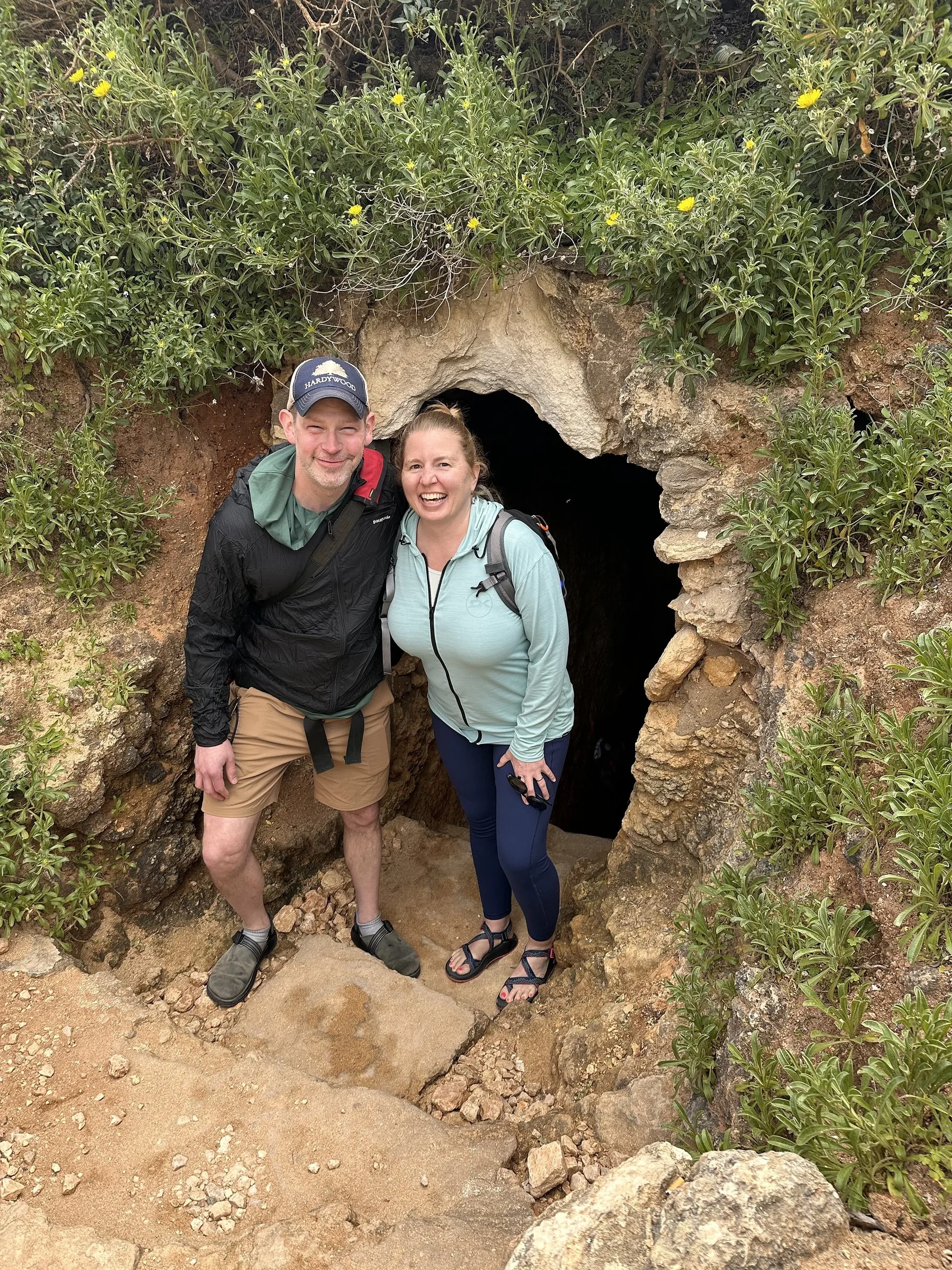 Smiling man and woman in hiking clothes standing in a small cave entrance framed by wildflowers