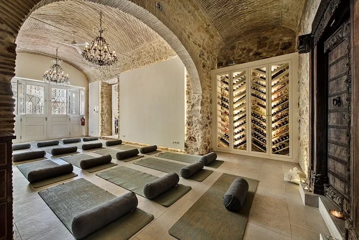 Yoga mats and bolsters in rows under vaulted stone arches and chandeliers, beside a lit wall of wine bottles