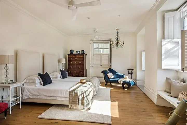 Sunny bedroom with two twin beds, wood floors, a crystal chandelier, and a blue chaise lounge by the window