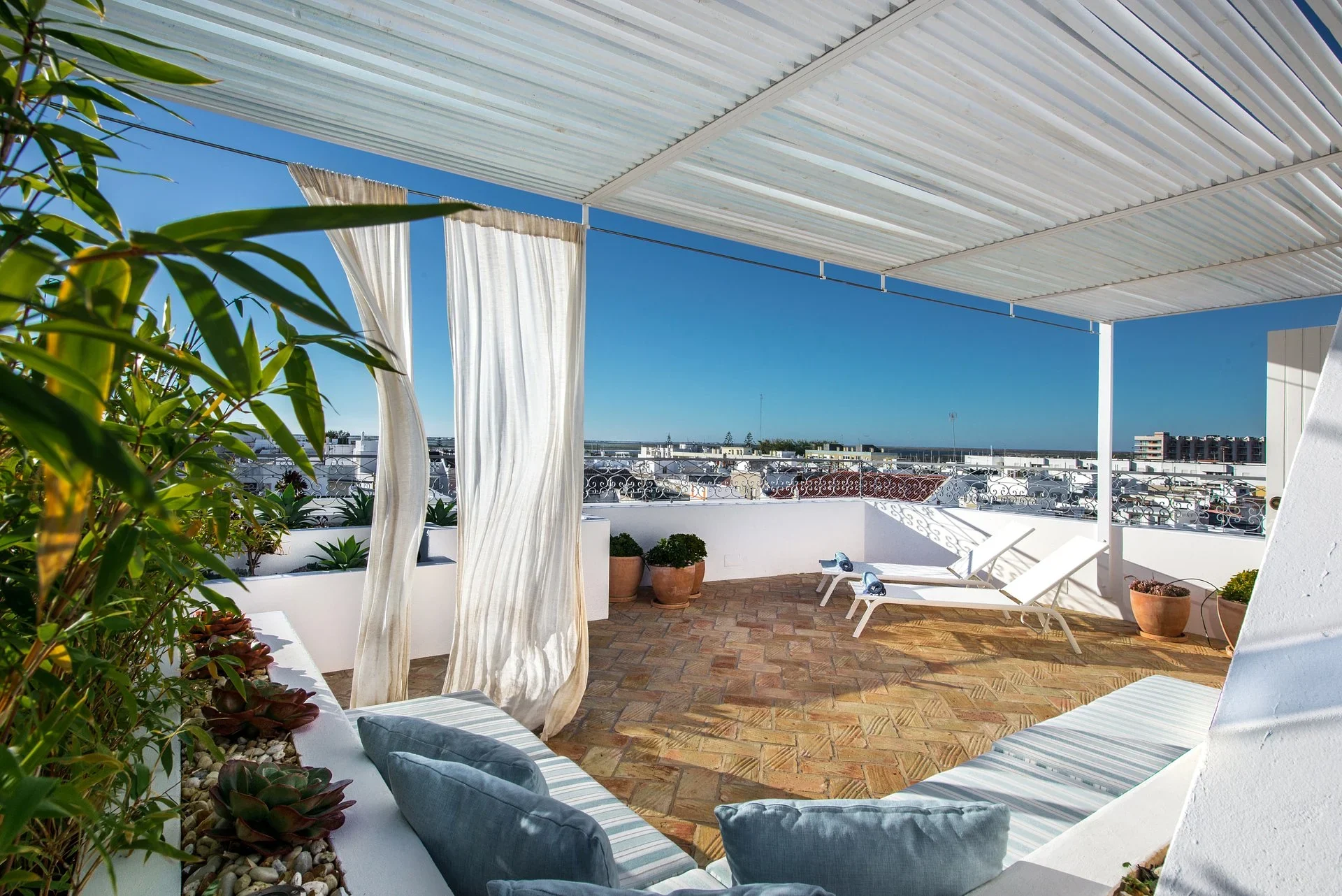 Private rooftop terrace with a slatted white pergola, flowing curtains, cushioned benches, and loungers over the town