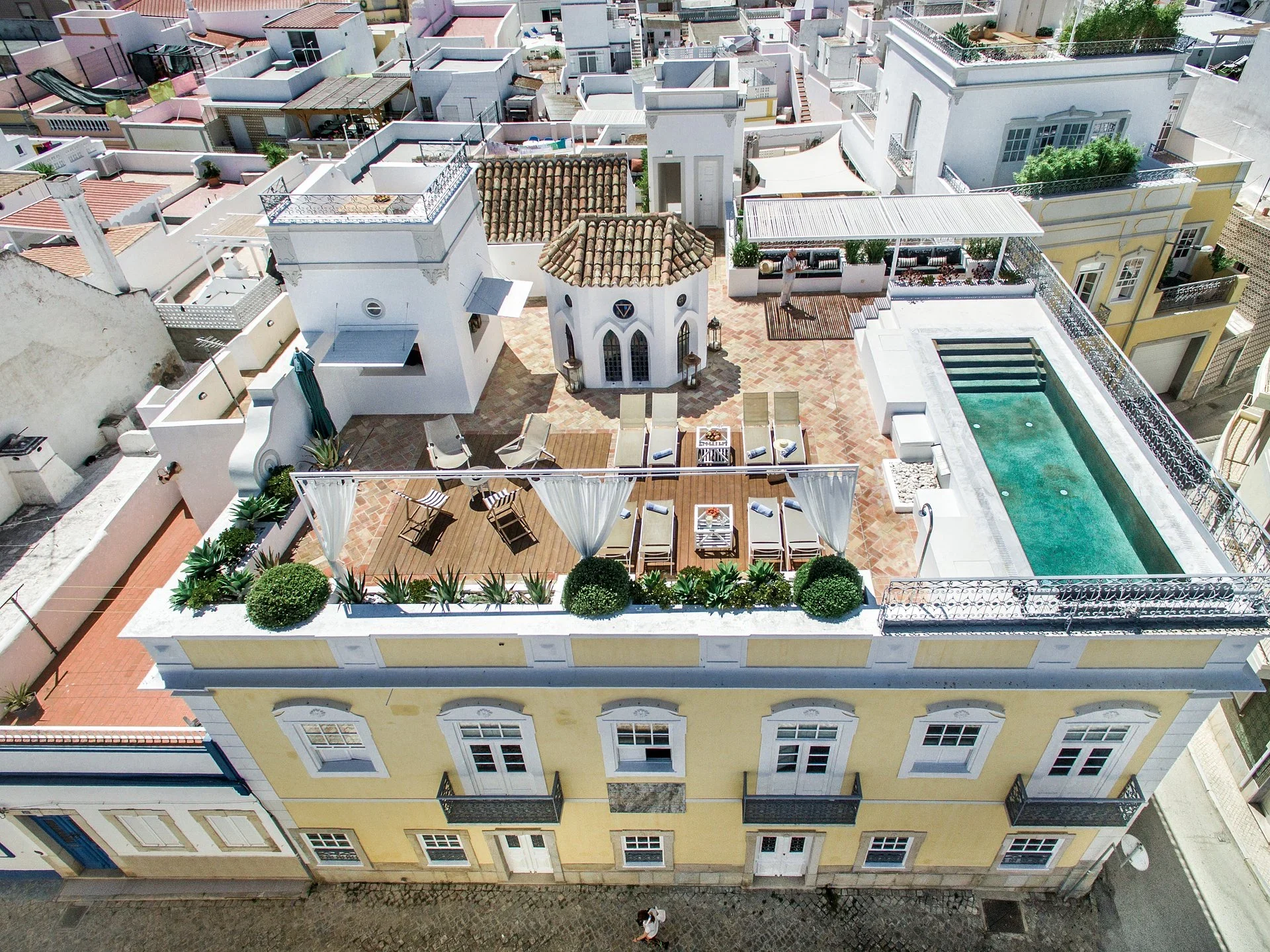 Aerial view of a yellow historic house with a rooftop terrace, sun loungers, a white chapel, and a long pool
