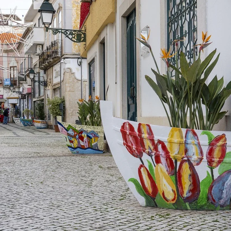 Old boats painted with tulips and colorful houses used as planters along a cobblestone shopping street