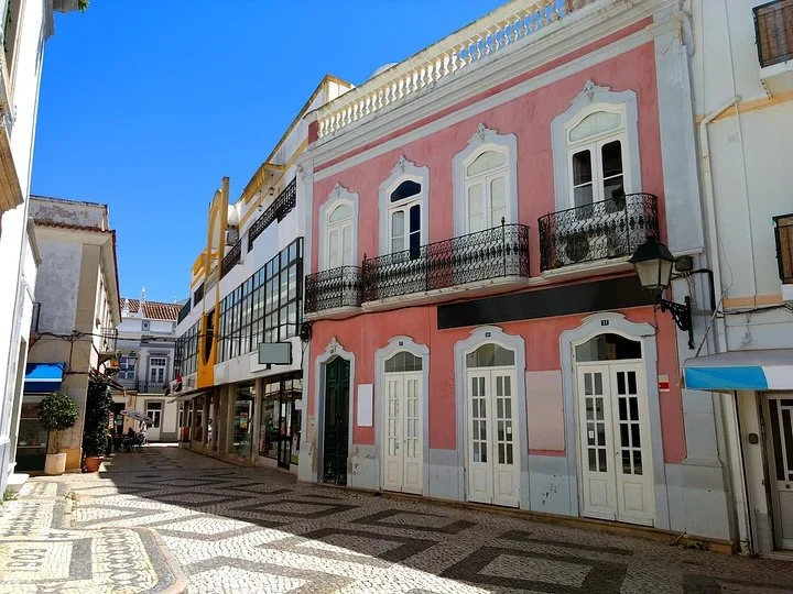 Pink two-story building with white-framed doors and iron balconies on a patterned cobblestone street