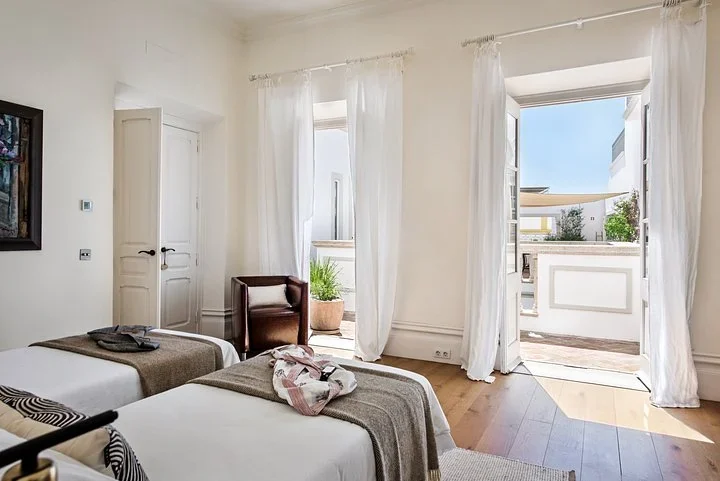 Bedroom with white linen curtains and tall doors open to a small balcony above the sunny courtyard