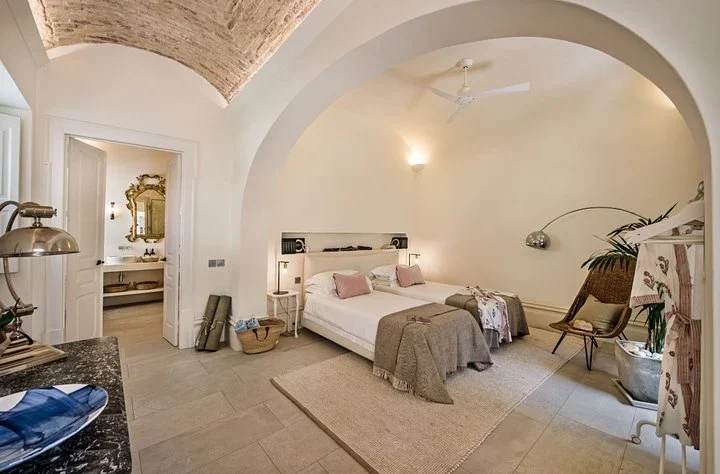Twin beds with pink pillows under a vaulted brick ceiling and a white arch, with an en-suite bathroom beyond
