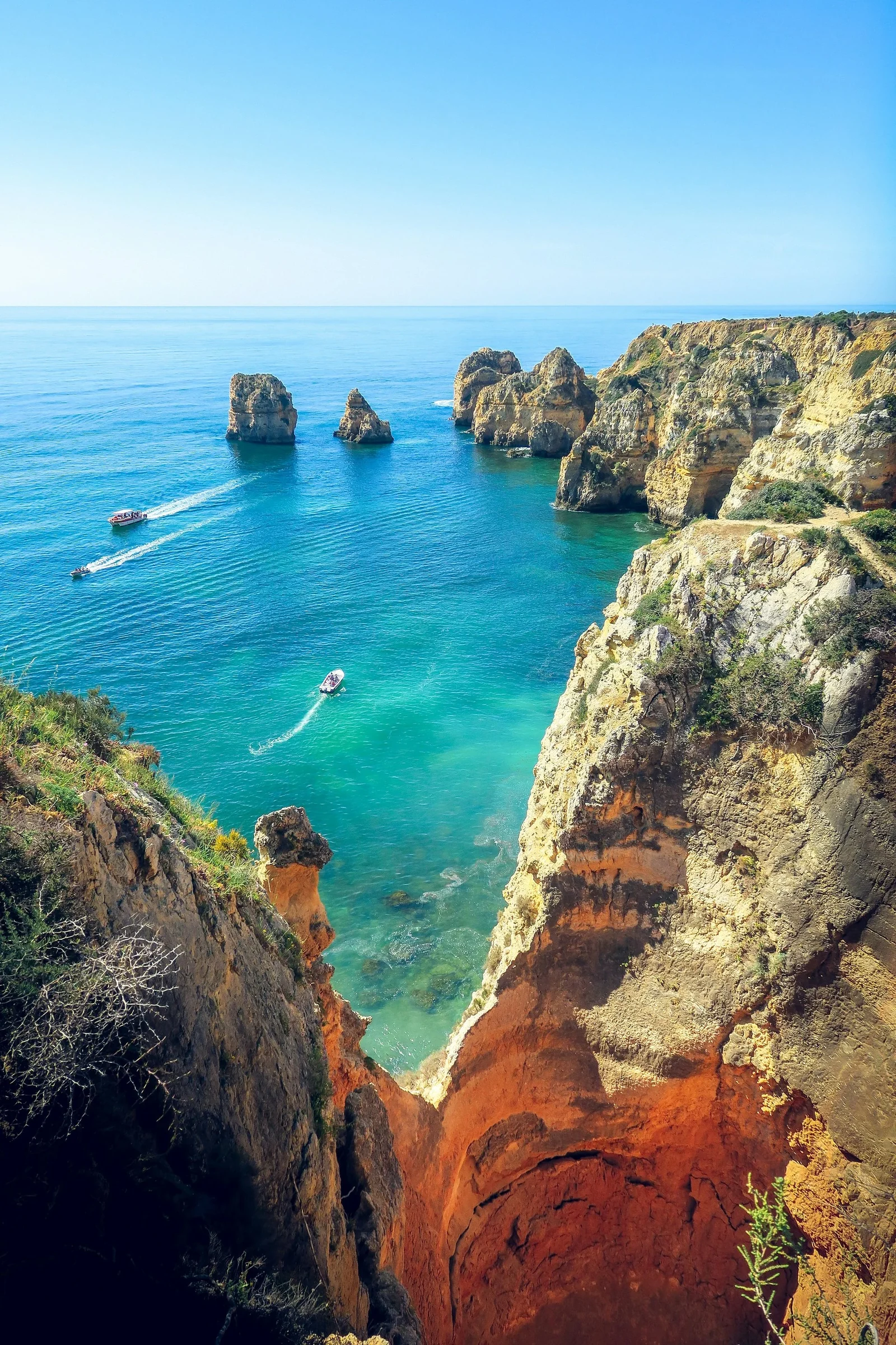 Boats cruising past sea stacks in turquoise water below tall golden cliffs on the Algarve coast