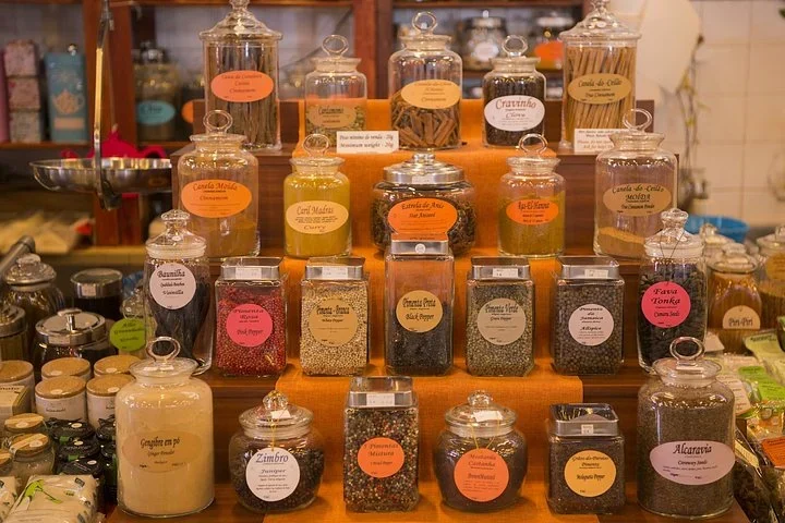 Glass jars of spices with handwritten Portuguese labels stacked on an orange display in a shop
