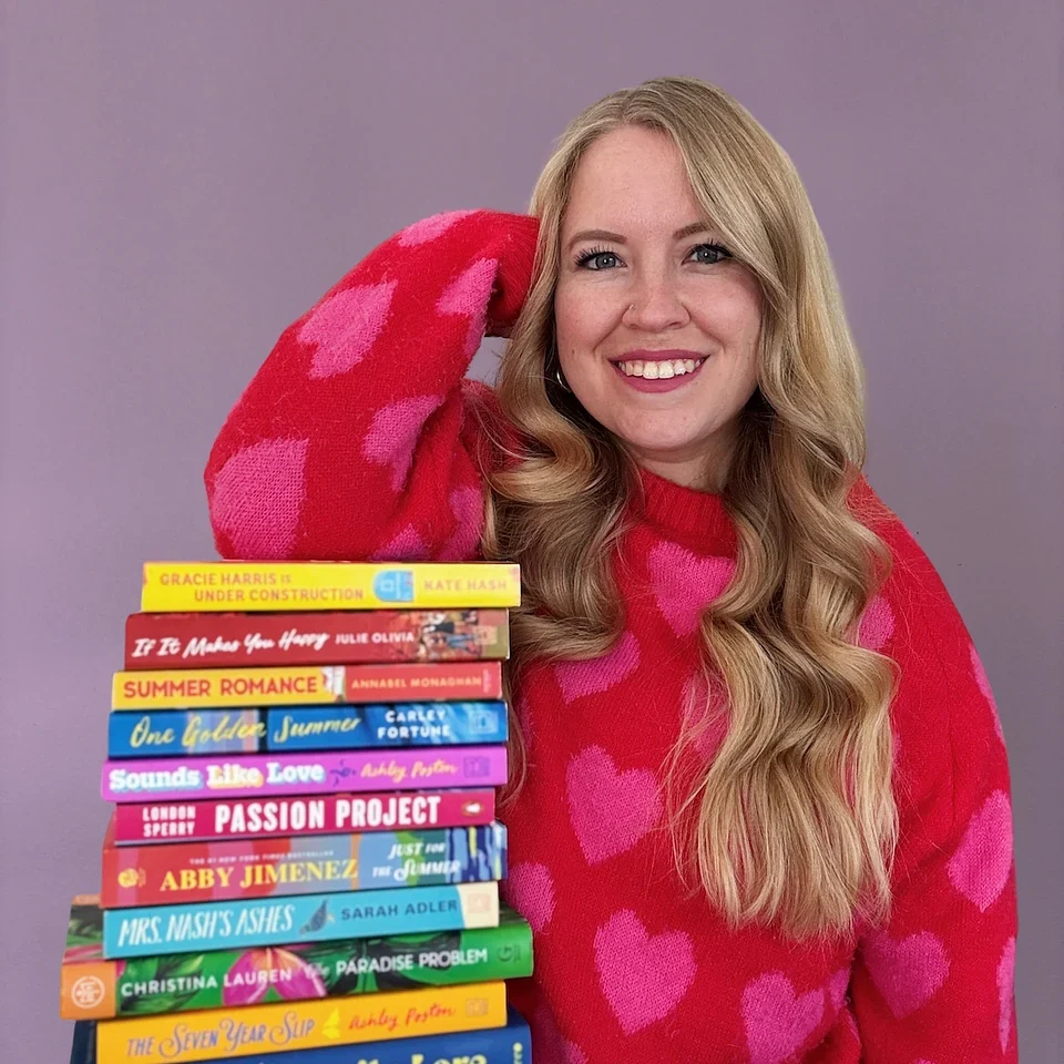 Anna from AllTooWellRead smiling beside a tall stack of colorful books