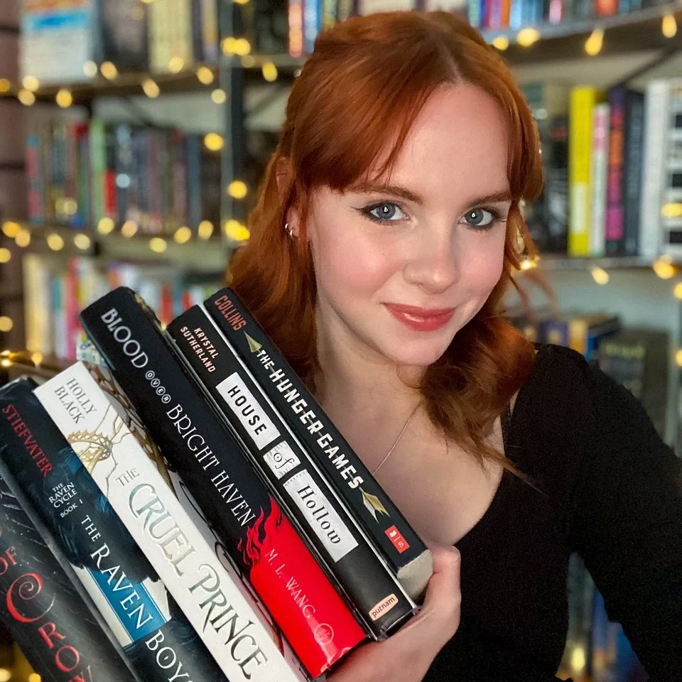 Daphne from daphsreads holding a stack of books in front of lit bookshelves