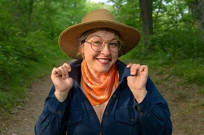 Anna Klimmek smiling in a straw hat, glasses, and an orange scarf on a wooded trail