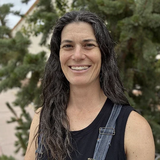 Deb Bass O'Brien smiling outdoors in front of evergreen trees