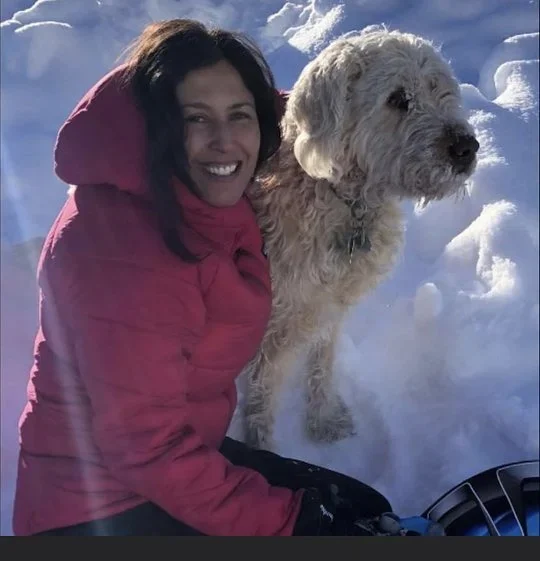 Dr. Ronit Berdugo smiling in a red winter jacket beside a shaggy white dog in the snow