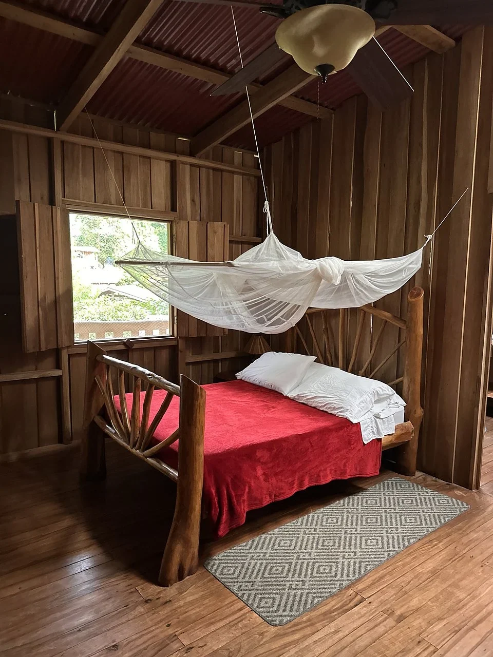 Log bed with a red blanket and mosquito net in a wooden cabin bedroom with a ceiling fan