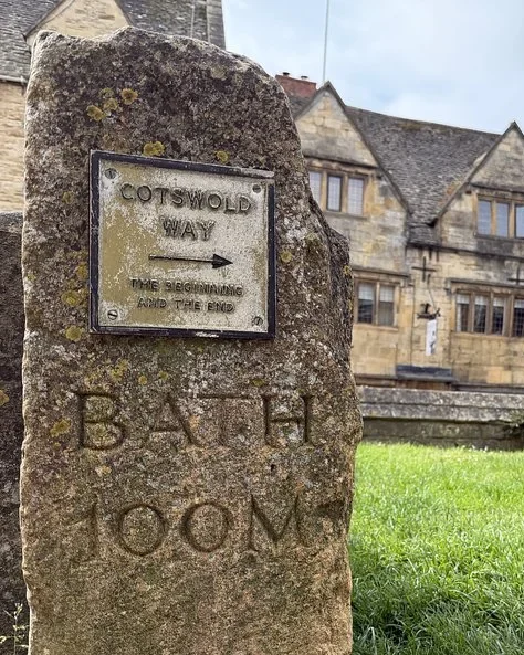 Stone Cotswold Way marker carved with Bath 100 miles in front of golden stone buildings