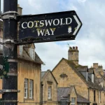 Black Cotswold Way footpath sign with an acorn symbol in front of golden stone buildings