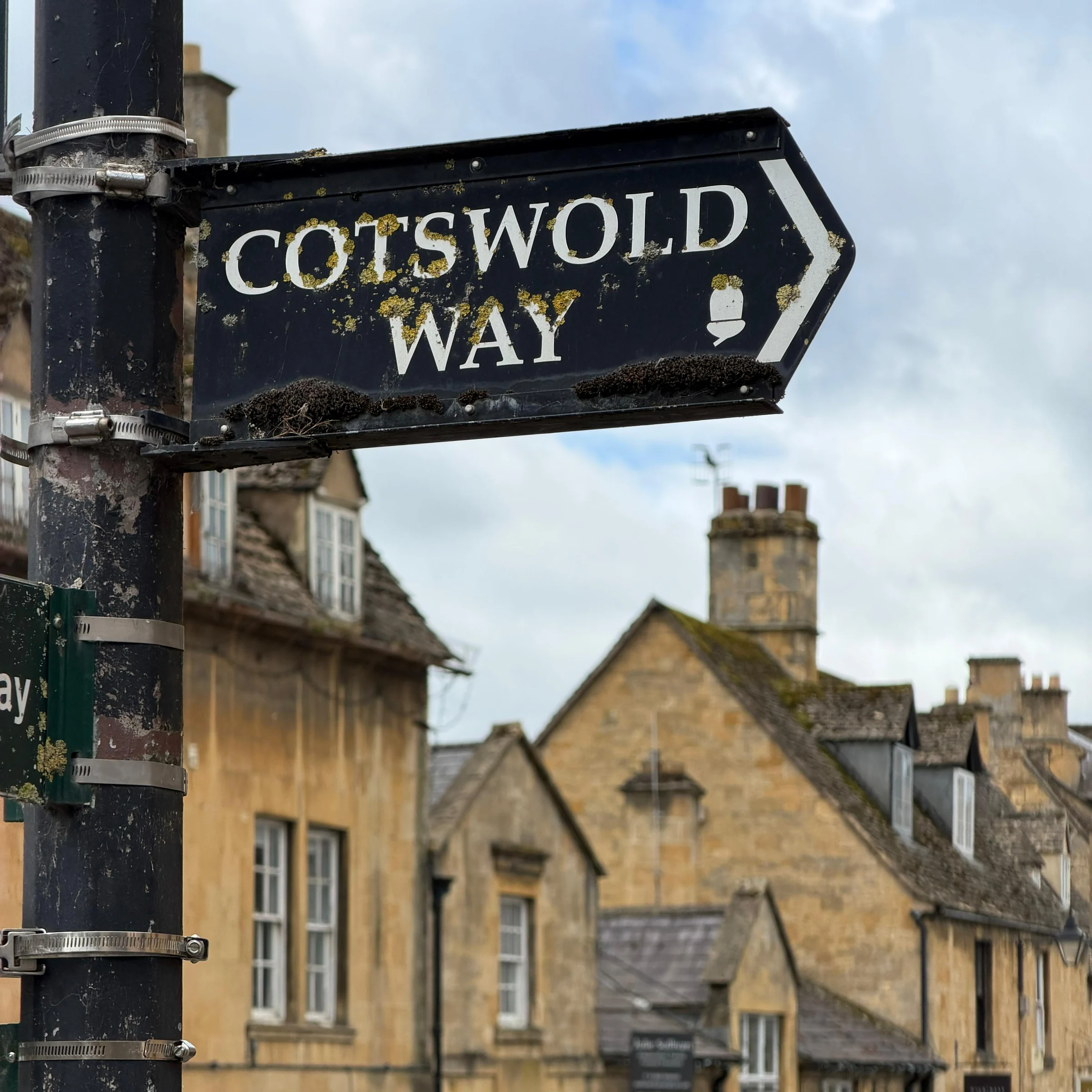 Black Cotswold Way footpath sign with an acorn symbol in front of golden stone buildings
