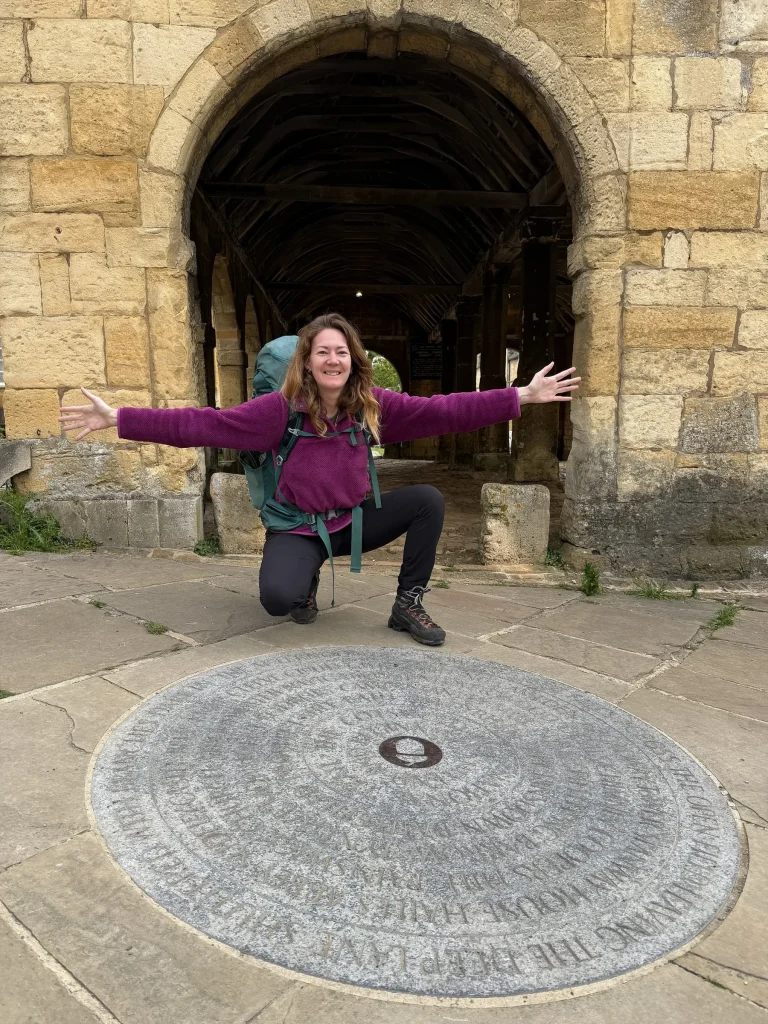 Smiling hiker with arms outstretched beside the round Cotswold Way stone marker under a stone market hall arch