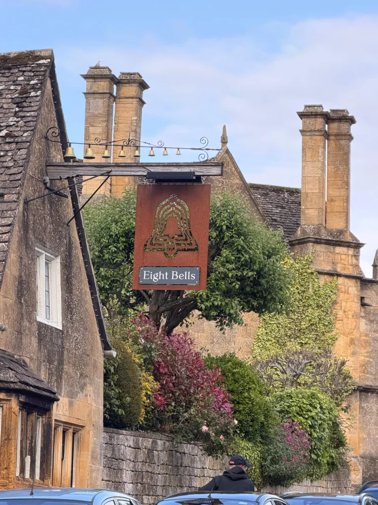 Eight Bells pub sign hanging among golden stone chimneys and flowering shrubs