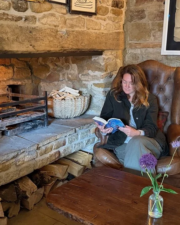 Woman reading a book in a leather armchair beside a crackling stone fireplace