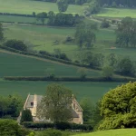 Sheep and lambs resting on a green hillside above a stone manor house and patchwork fields
