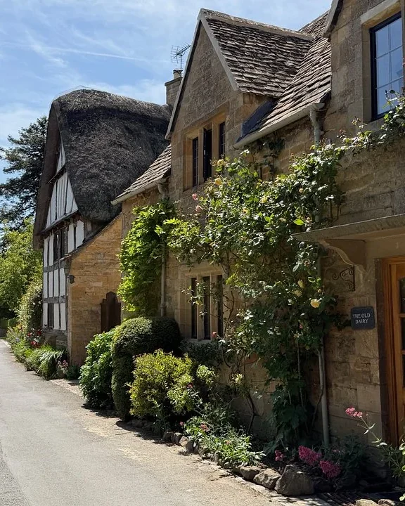 Thatched cottage and rose-covered stone houses along a quiet Cotswold village lane