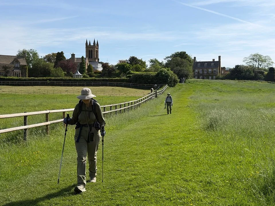 Walkers with trekking poles on a mown grass footpath beside a fence, with a church tower and stone houses behind