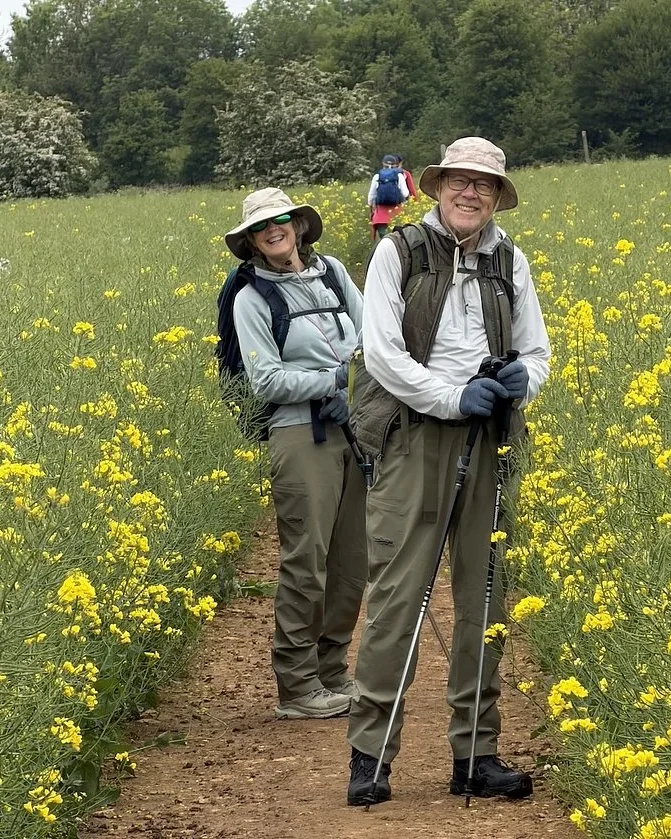 Two smiling walkers with light day packs on a path through a field of yellow flowers
