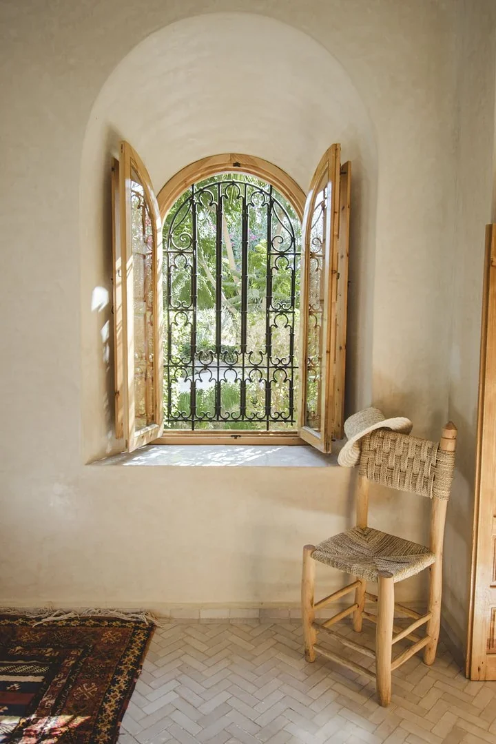 Arched window with an iron grille and a woven chair holding a straw hat