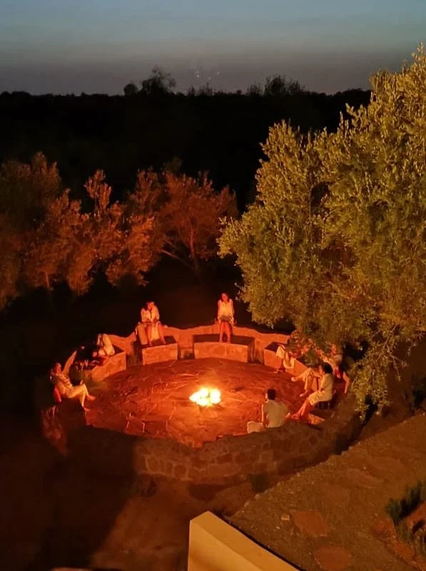 Guests gathered around a stone fire pit at dusk, framed by olive trees