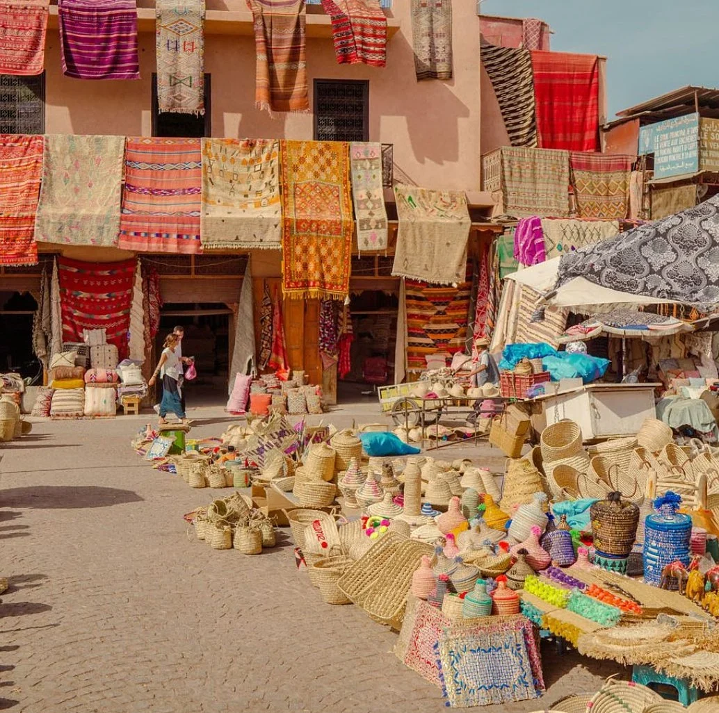 Market square lined with hanging rugs, woven baskets, and colorful straw containers