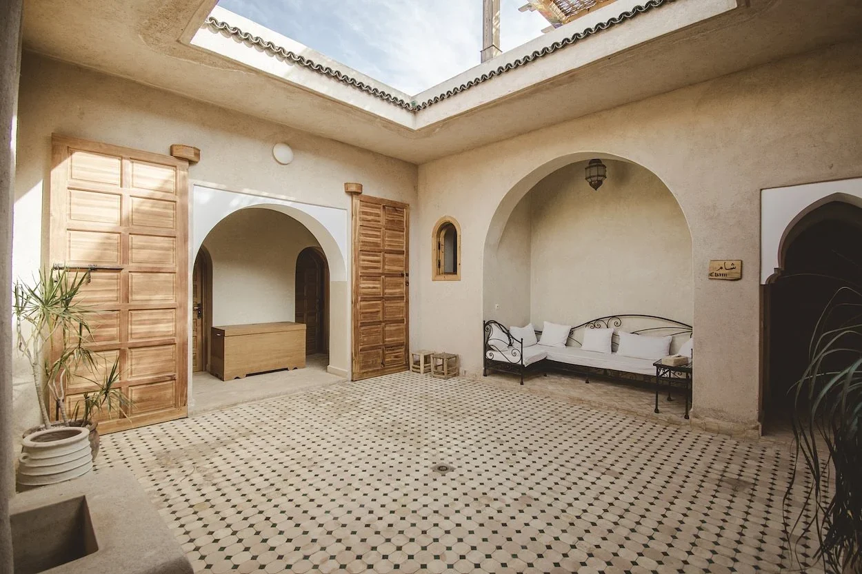 Open-roof riad courtyard with a zellige tile floor, carved wooden doors, and a cushioned daybed in an arched nook