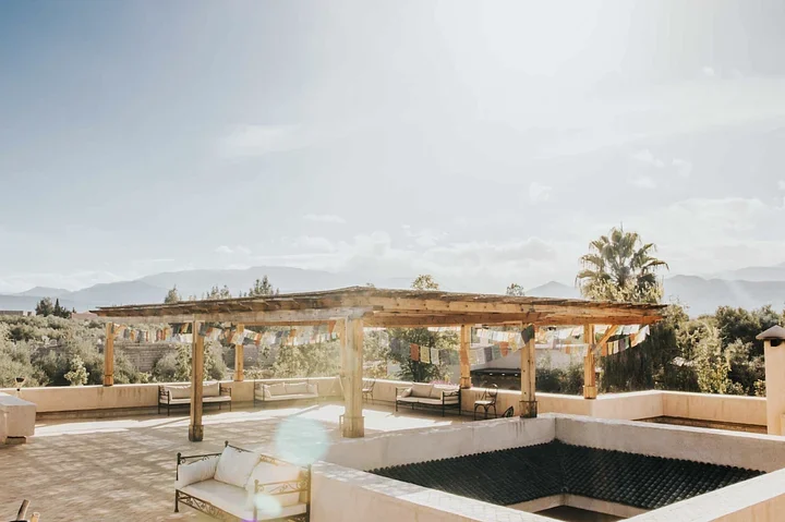 Sunny rooftop terrace with daybeds under a wooden pergola and prayer flags, mountains in the distance
