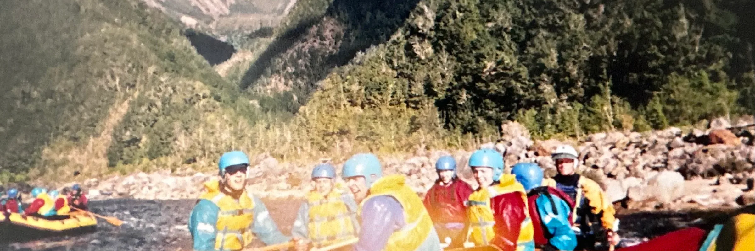 Group in blue helmets and life jackets in a yellow raft on a river below forested mountains in New Zealand