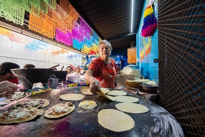 Oaxaca culinary retreat stop at a market stall where a cook presses memelas on a large comal under papel picado