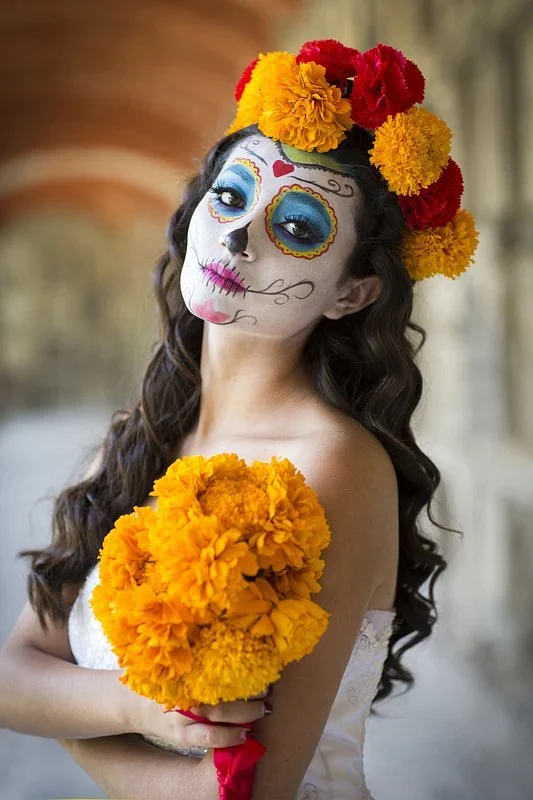 Woman with Catrina face paint and a crown of marigolds and red flowers, holding a bouquet of marigolds under a stone arcade