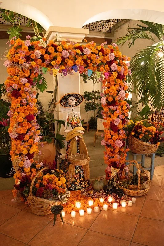 Arch of marigolds and cockscomb over a Catrina figure, woven baskets, and candles, set up as a Day of the Dead altar in a tiled courtyard