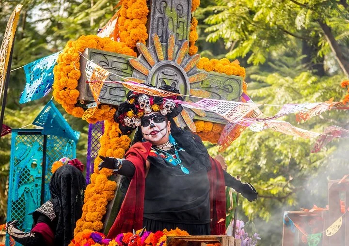 Woman dressed as La Catrina on a parade float covered in marigolds and papel picado, with trees behind