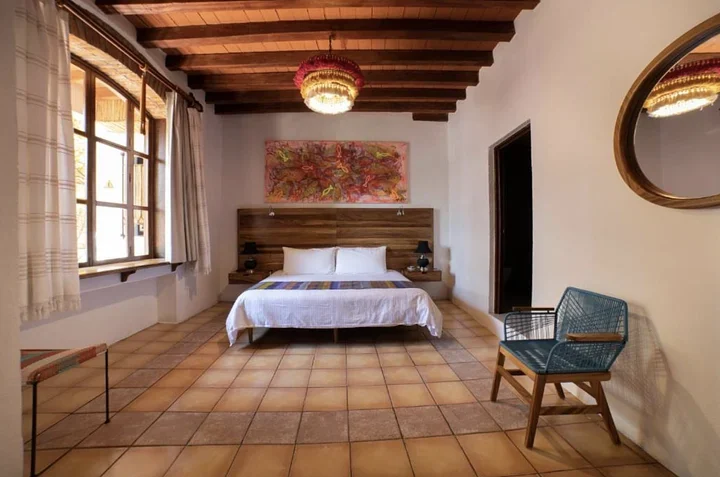 Hotel room with a king bed, a wood-beamed ceiling, terracotta floor tiles, and a blue woven chair
