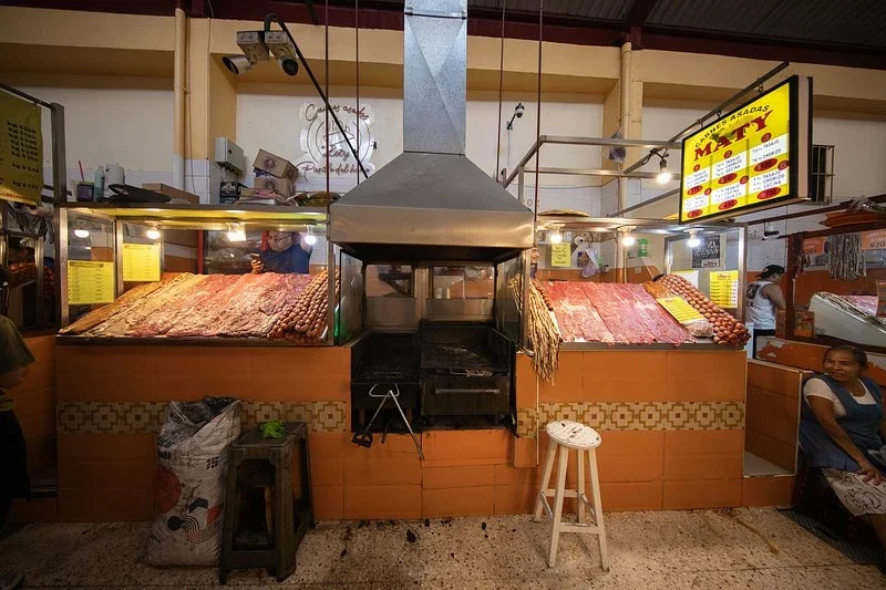 Market grill stall in Oaxaca with meats and chorizo laid out beside an open grill under a steel hood