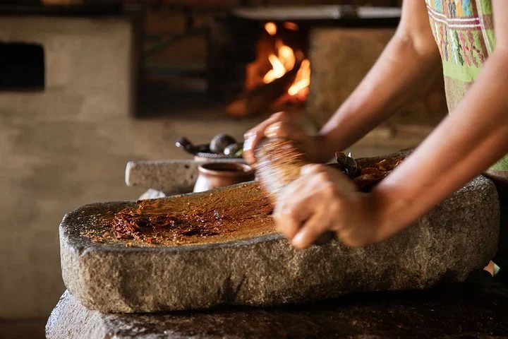 Hands grinding a paste on a stone metate in a traditional kitchen, with a wood fire burning behind