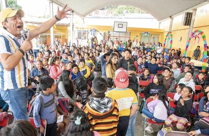 Room full of smiling students at a community event in Oaxaca, with a man speaking into a microphone