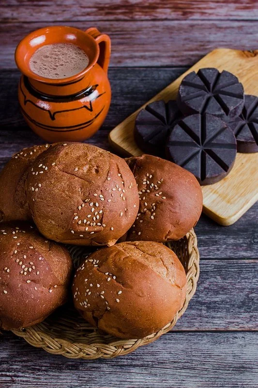 Basket of sesame-topped Mexican bread rolls beside tablets of Oaxacan chocolate and a clay jug of hot chocolate