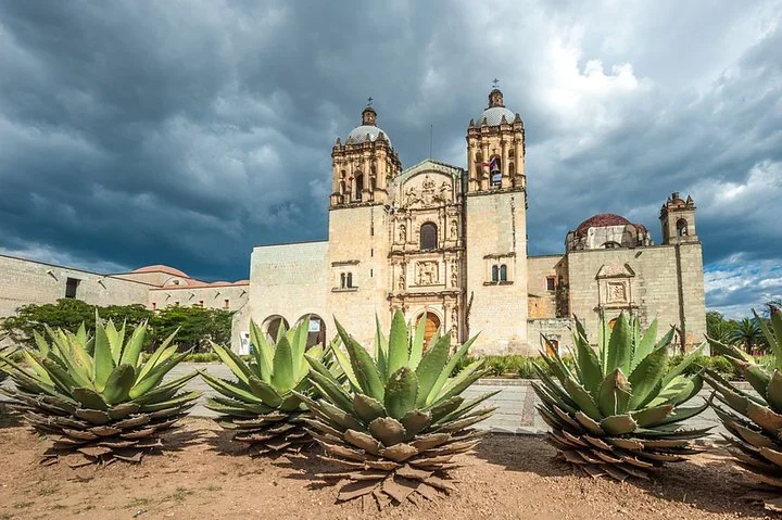 Templo de Santo Domingo de Guzm&aacute;n in Oaxaca under dramatic clouds, with large agave plants in front