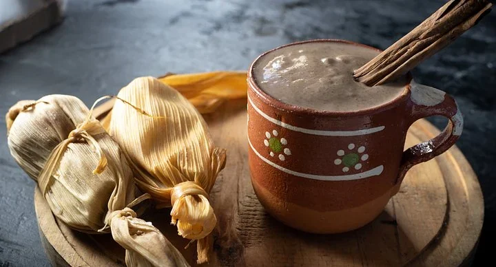 Tamales wrapped in corn husks beside a clay mug of champurrado with a cinnamon stick