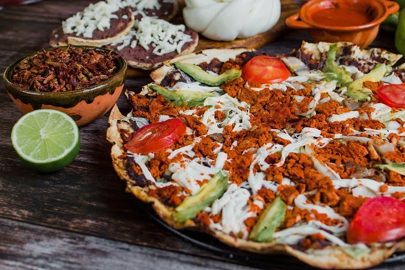 Crisp Oaxacan tlayudas topped with chorizo, quesillo, avocado, and tomato, with a bowl of chapulines and a lime