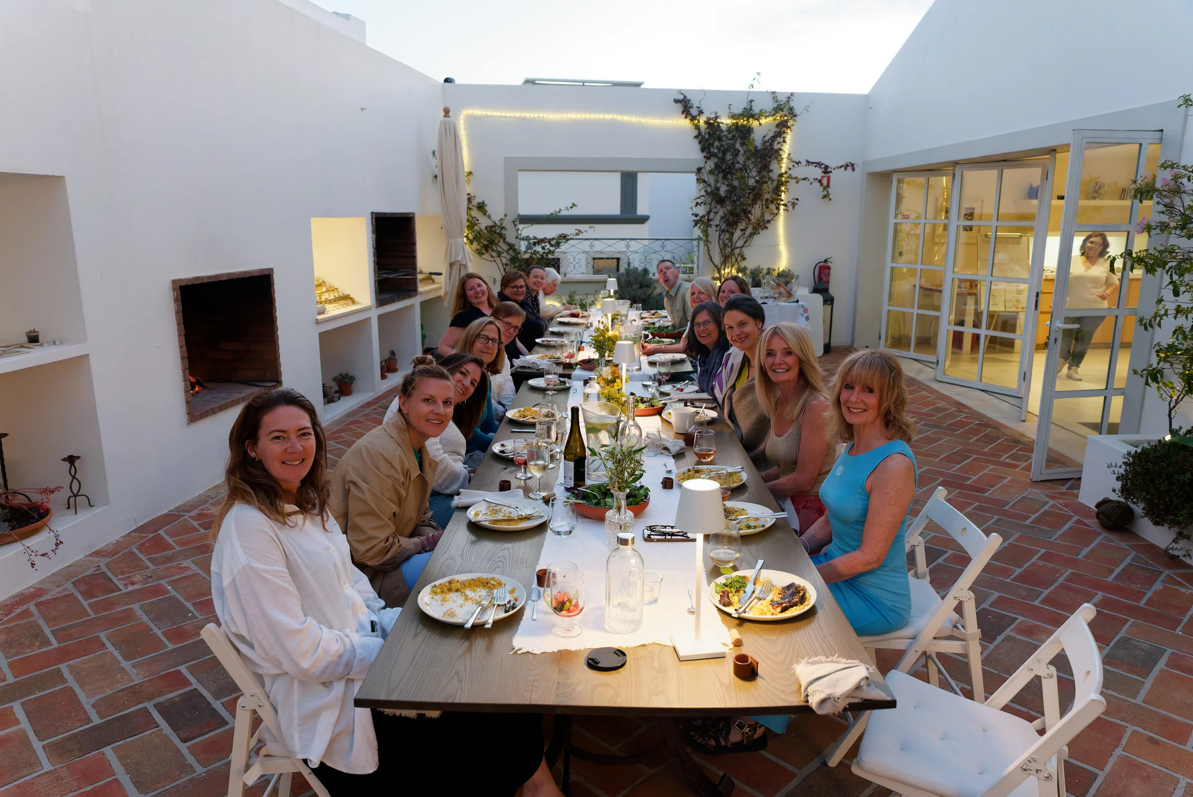Retreat guests smiling around a long candlelit table in a whitewashed courtyard at dusk